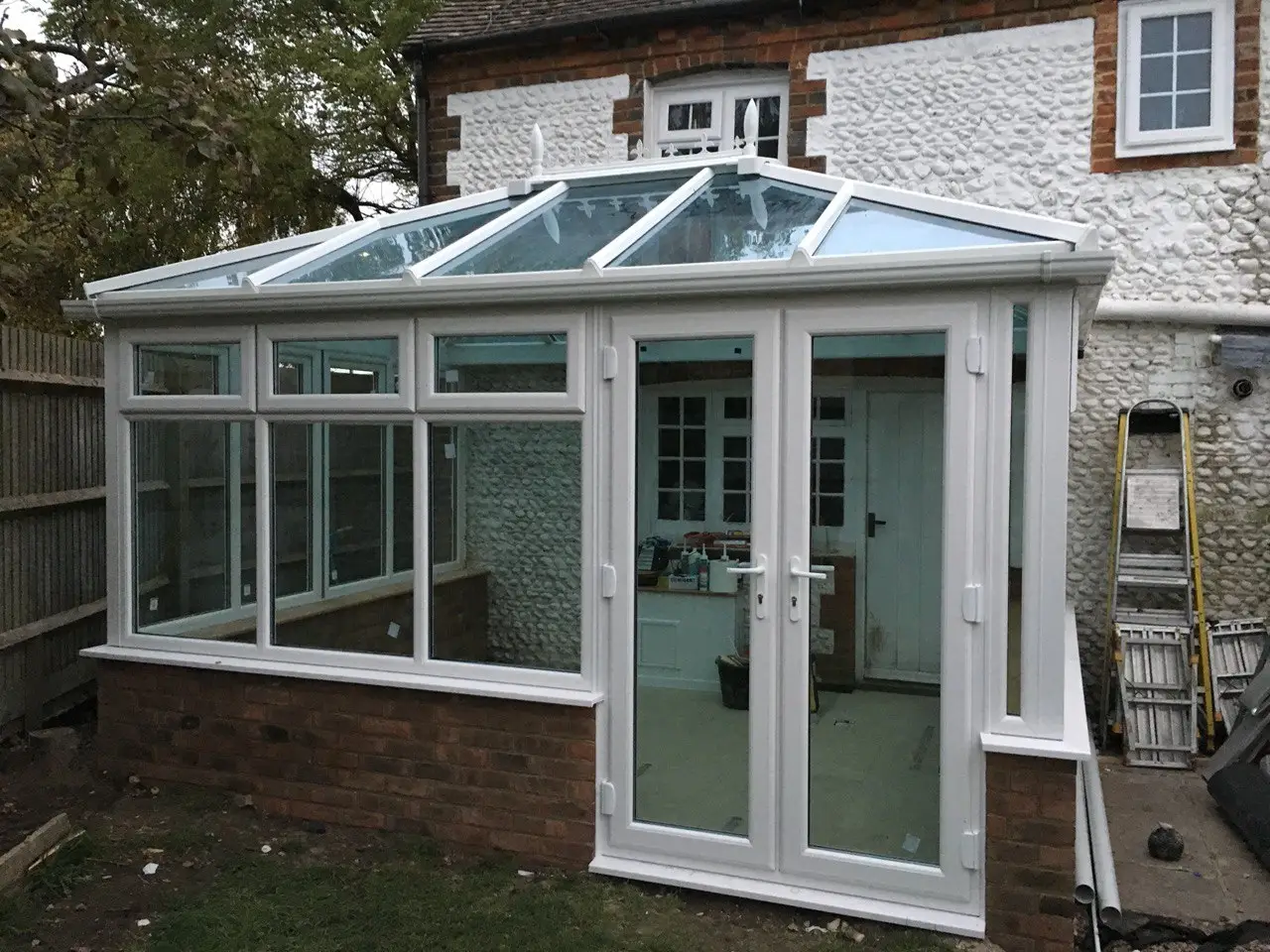 A white conservatory with a glass roof and French doors, built onto a flint and brick house