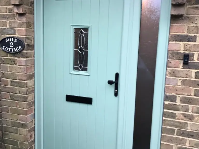 A sage green composite front door with a patterned glass panel set in a brick porch